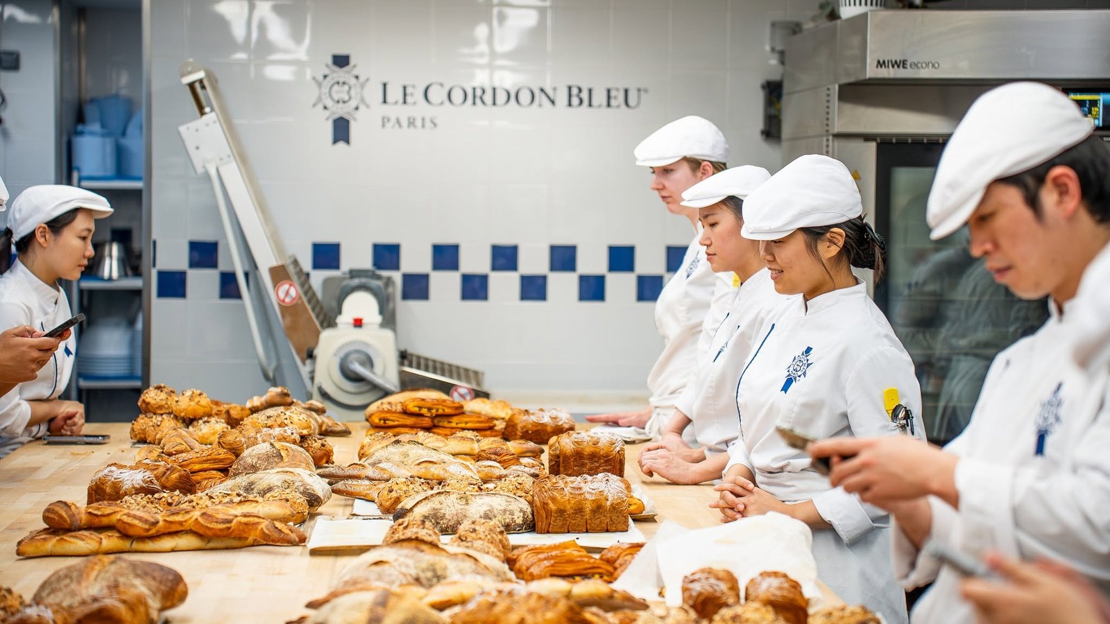 Le Cordon Bleu Paris 麵包工房：學生列隊評審成品，工作檯鋪滿可頌、丹麥酥及歐式麵包，牆上為巴黎校區標誌